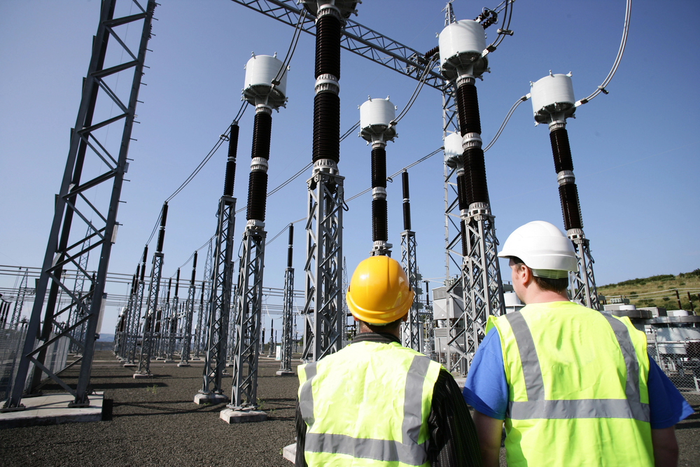 Two utility workers in hard hats and safety vests walking through a high-voltage substation